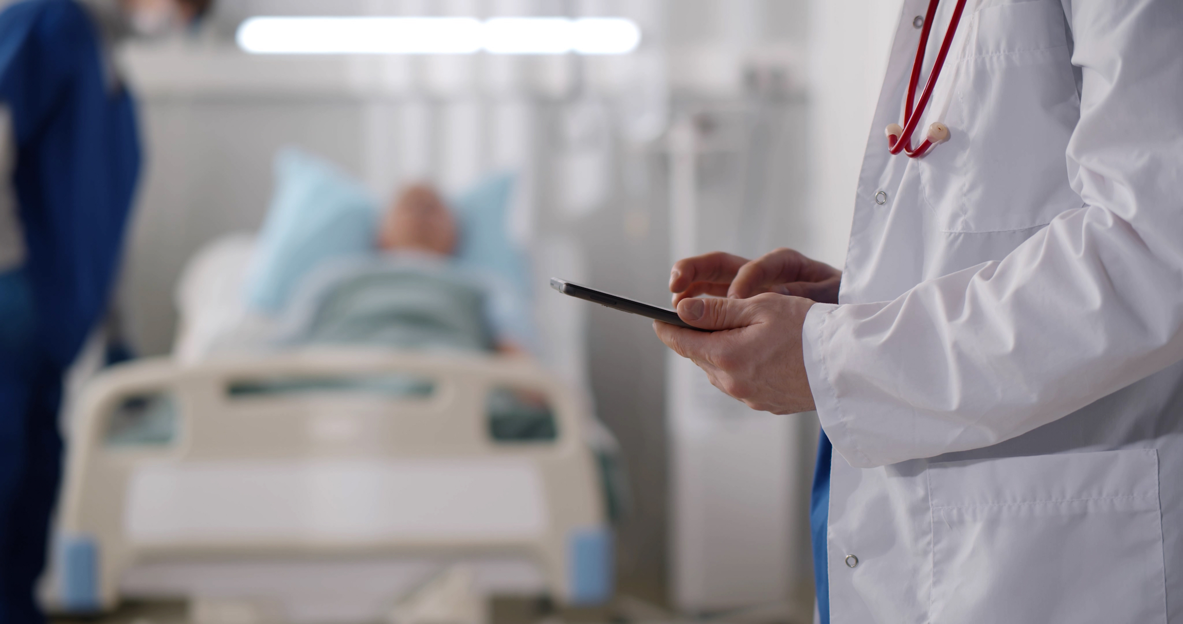 Przycięty strzał lekarza w mundurze ze stetoskopem przy użyciu tabletu PC stojącego na oddziale szpitalnym z pacjentem spoczywa w łóżku na tle. Praktyk sprawdzający dane pacjenta na tablecie cyfrowym