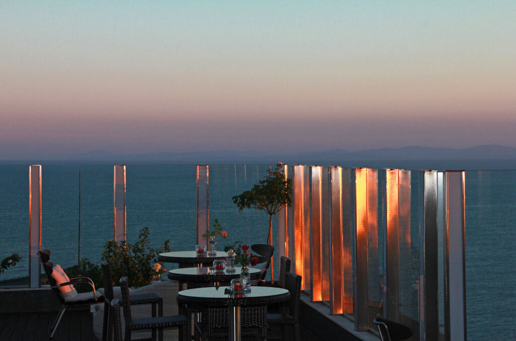Kawiarnia zewnętrzna na dachu hotelu ze stolikami i krzesłami rattanowymi, z panoramicznym widokiem na morze z odblaskami słońca na szklanym ogrodzeniu o zachodzie słońca. Morze Czarne, Bułgaria, miejscowość Ravda, aparthotel Emerald.