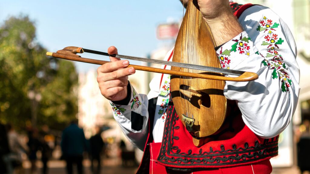 Bulgarian folk musician - violinist in traditional national costume plays an old stringed instrument - gadulka Blurred city street in background. Plovdiv Bulgaria. Bulgarian folklore and culture